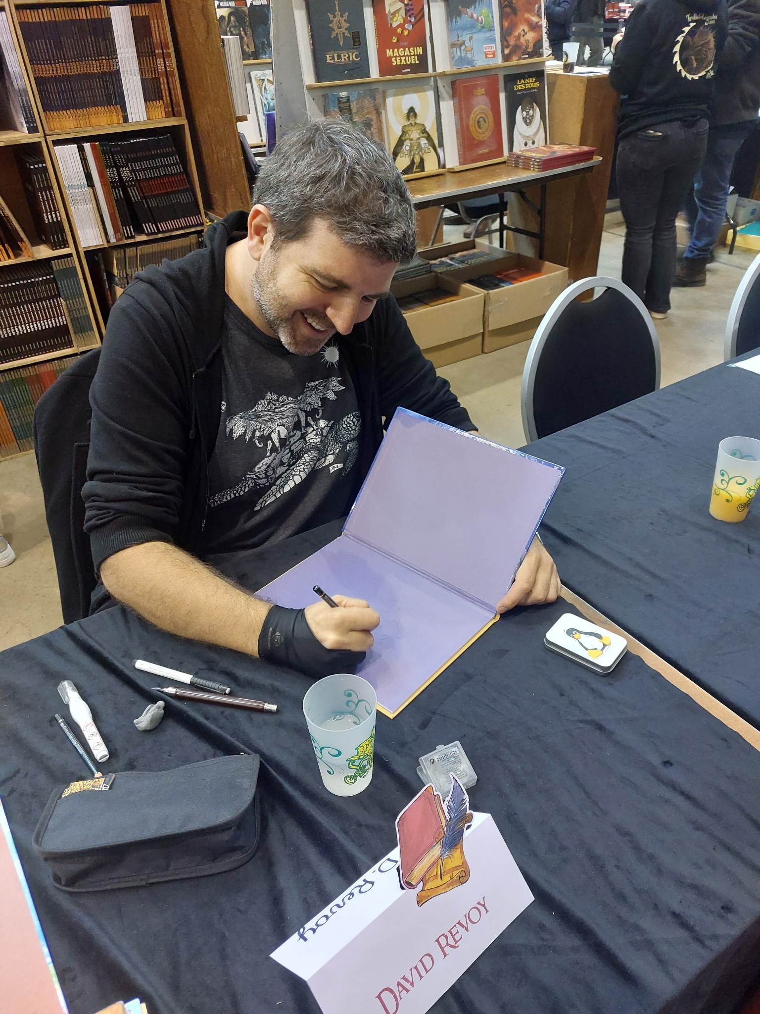 A photo of David Revoy, photo made from the point of view of a visitor at the start of having the book four of Pepper&Carrot signed at Trolls & Legendes Festival, in Mons Belgium, 2023. It's a top-down photograph in color made from a smartphone of a seated white person around 40 years old smiling and happy, with a short beard and a mix of brown and white hair. He wears a tee shirt with the great Great A'Tuin (Discworld, T.Pratchett), and also wears a glove for drawing (synthetic, black) and starts to doodle into a book open. The book is Pepper&Carrot four by Glénat, with its dark violet colors in the introduction. The table has drawing material on it (Tipp-Ex, Pierre Noire, white markers, and a kneaded eraser), a little box with a tux sticker on it, a glass of water, and the table are covered with fabric in black. A label "David Revoy" is on the front to indicate the booth to the visitor. In the background: the library shop of the Festival with many comic books. Photo CC-By 4.0 Steph Vesch.